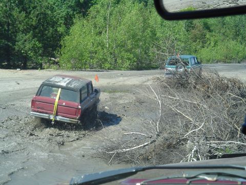 1993 Ford Bronco - Two SUVs navigate a muddy off-road course with dense trees surrounding the area.