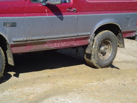 1993 Ford Bronco - The side of a red and silver Ford Bronco is shown on a dirt surface, with mud on the tires and lower body panels.