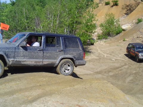 1993 Ford Bronco - A dark blue Jeep Cherokee XJ navigates a challenging off-road course with another vehicle visible in the background.