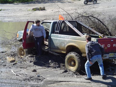1993 Ford Bronco - A muddy Ford pickup truck is stuck in a watery, debris-filled area with two men nearby.