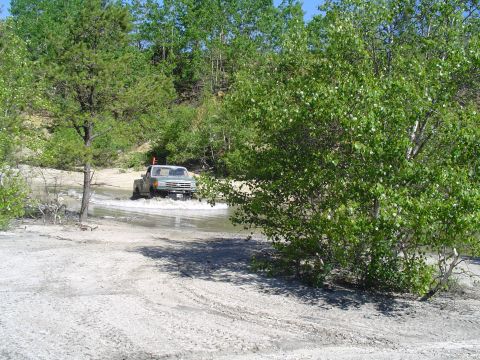 1993 Ford Bronco - A pickup truck navigates a water crossing amidst trees and sandy terrain.