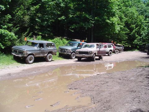 1993 Ford Bronco - Several SUVs are parked on a dirt road next to a forest, with a large puddle in the foreground.