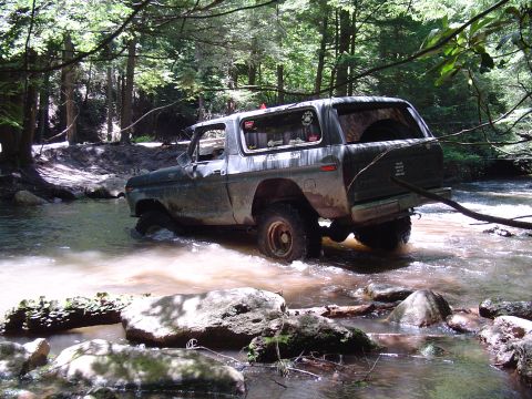 1993 Ford Bronco - Older model Ford Bronco navigates a rocky stream bed surrounded by trees.