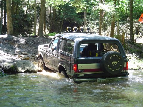 1993 Ford Bronco - A Ford Bronco navigates a rocky stream bed surrounded by trees.