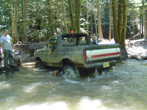 1993 Ford Bronco - A 1993 Ford Bronco navigates a water obstacle in a forest setting, kicking up spray.