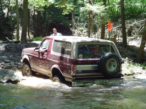1993 Ford Bronco - A red and tan 1993 Ford Bronco navigates a rocky stream in a wooded area.