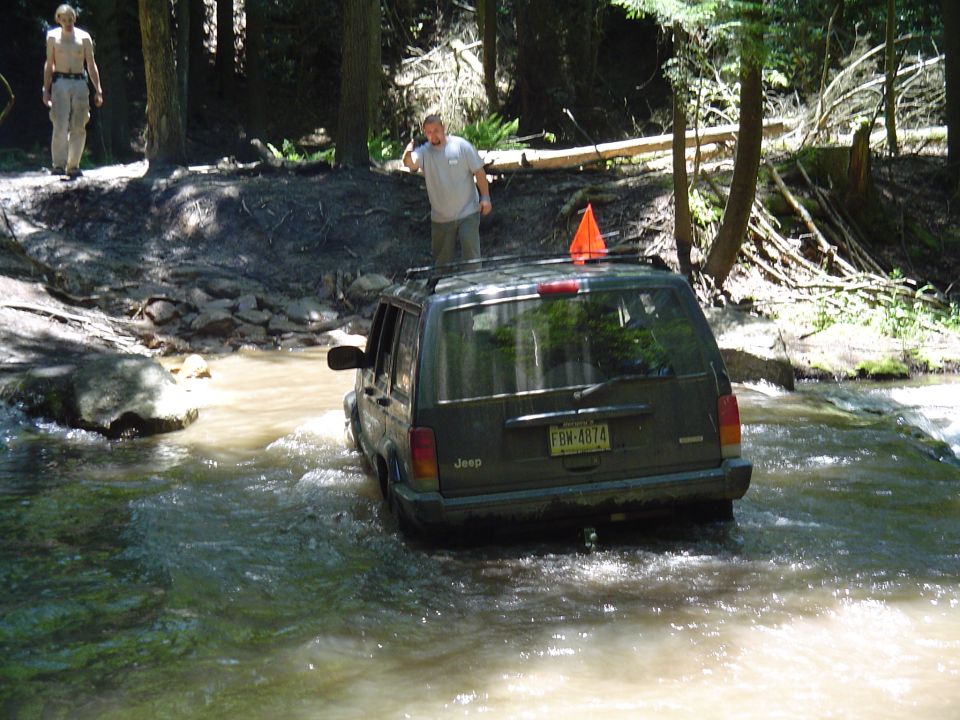 Jeep Cherokee XJ driving through a shallow stream in a wooded area. Two men are visible on the bank