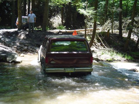1993 Ford Bronco - A maroon Ford Bronco navigates a rocky stream in a wooded area, with another vehicle and a person visible in the background.