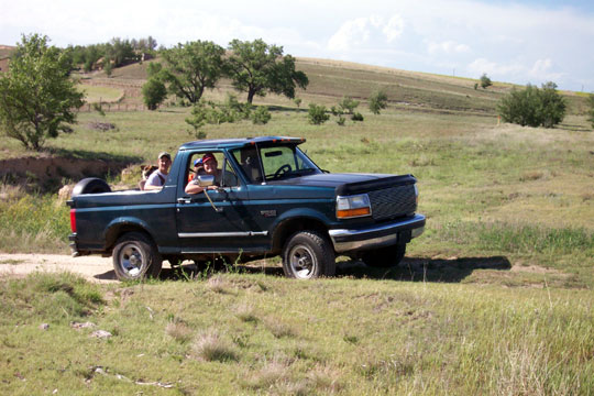 Dark green 1995 Ford Bronco with people in the cab and bed, driving on a dirt road