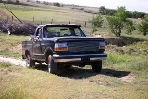 1995 Ford Bronco - A dark green Ford pickup truck drives down a dirt path through a rural, grassy landscape.