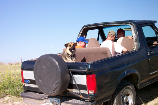 Dark Ford Bronco with top removed, dog and people inside in open field