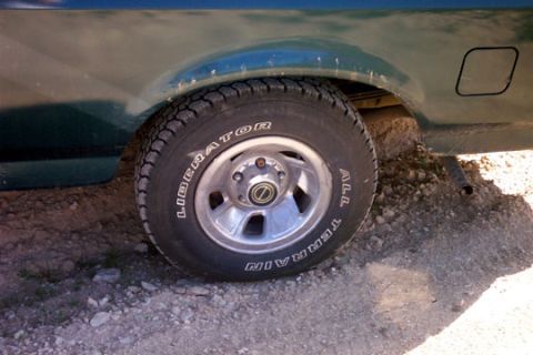 1995 Ford Bronco - Close-up of the rear wheel of a dark green truck parked on gravel, fitted with a raised-white-letter Liberator All Terrain tire on a five-l…