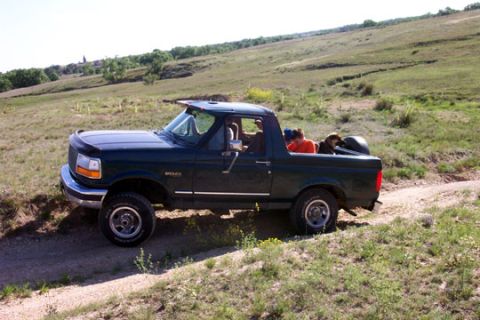 1995 Ford Bronco - Dark green 1990s Ford Bronco with its rear top removed, parked at a front-left three-quarter angle on a dirt two-track through open grassla…