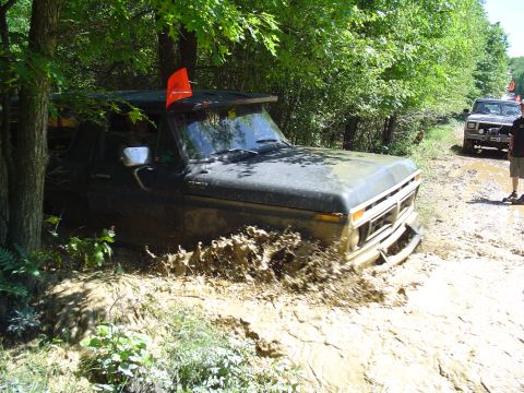 1993 Ford Bronco - A muddy Ford F-Series truck is stuck in a dirt path surrounded by trees, with another vehicle and a person in the background.
