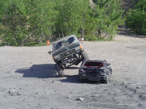 1993 Ford Bronco - A green Ford truck is positioned precariously on a damaged black car in a dirt lot.