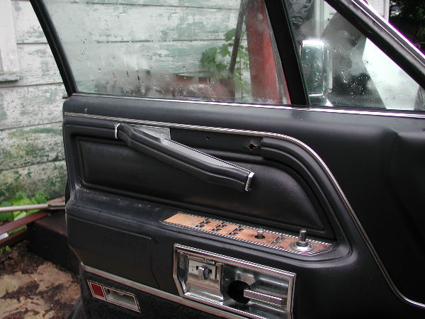 Black car door panel with woodgrain power window switch plate and missing interior door handle