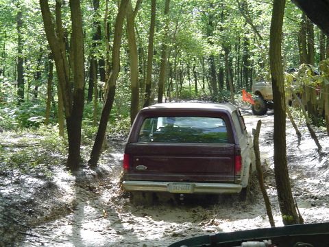 1993 Ford Bronco - A maroon 1993 Ford Bronco navigates a muddy trail in a wooded area, with another off-road vehicle visible in the background.