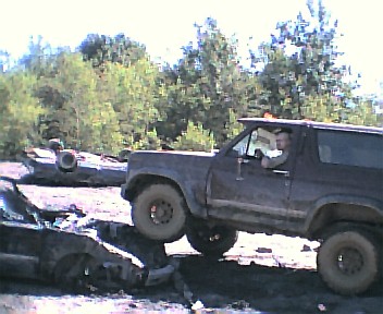 1985 Ford Bronco - A Ford Bronco navigates over a pile of wrecked vehicles in a junkyard or off-road area.