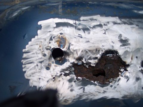1990 Ford Bronco - Close-up of rusted metal with grinding marks and a hole, indicating body work on a vehicle part.