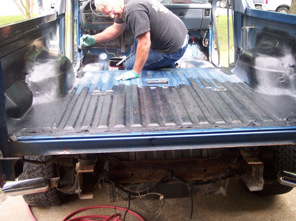 Man rolling black bedliner coating in stripped rear floor of blue Ford Bronco