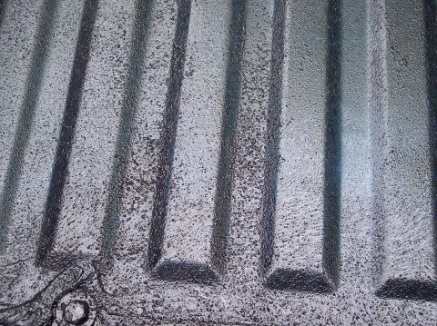 1990 Ford Bronco - Extreme close-up of a ribbed sheet-metal floor pan scuffed and abraded down to bare metal, with the stamped ridges showing dark residue and…