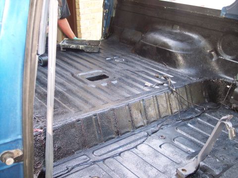1990 Ford Bronco - Close view of the bare rear floor and wheel tub of a blue SUV freshly coated in textured black roll-on bedliner, with the ribbed floor pan,…