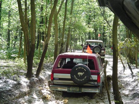 1993 Ford Bronco - Two Ford Broncos navigate a muddy path through a wooded area, kicking up dirt and water.