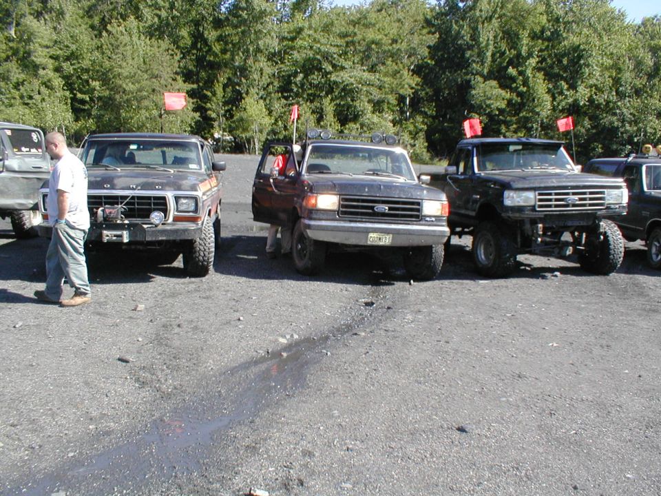 Three Ford trucks parked in a gravel lot. The leftmost truck is a dark color with a winch. The middle truck has its door open. The rightmost truck is lifted