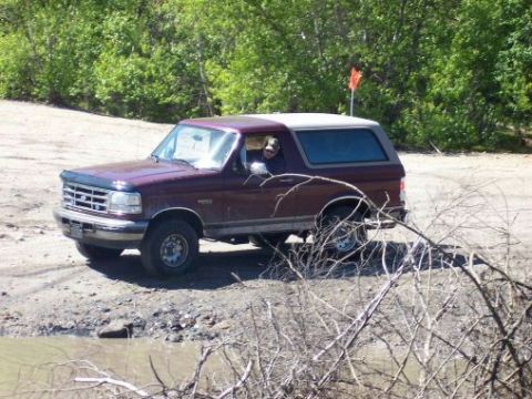 1996 Ford Bronco - A maroon 1996 Ford Bronco is seen parked on a dirt road with a dog visible in the driver's seat.