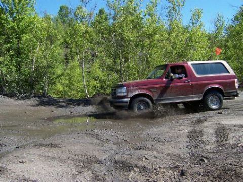 1996 Ford Bronco - A maroon 1996 Ford Bronco splashes through a muddy puddle on a dirt road, with trees in the background.