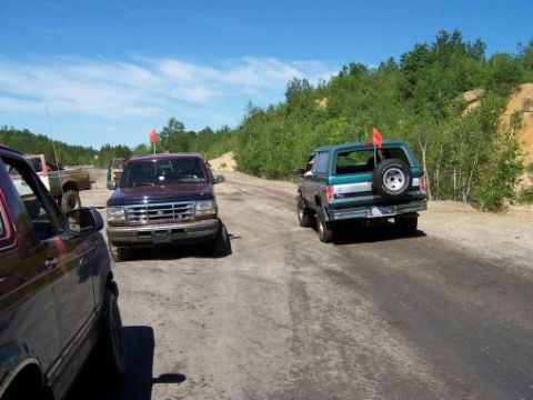 1996 Ford Bronco - Two Ford Broncos are seen driving on a dirt road under a bright blue sky.