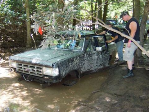 1996 Ford Bronco - A muddy black 1996 Ford Bronco is stuck in a muddy off-road course, with two people observing the situation.