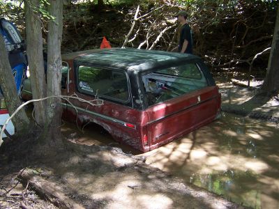 1996 Ford Bronco - A red SUV from the late 1970s is stuck in a muddy, wooded area with trees surrounding it.