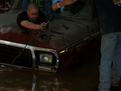 1996 Ford Bronco - A man is on the hood of a red pickup truck that is submerged in mud, with another person pulling on a rope attached rope.