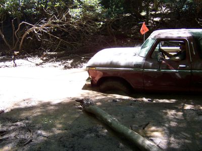 1996 Ford Bronco - A red Ford Bronco is stuck in the mud, with a driver inside and an orange flag on the roof.