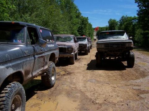 1996 Ford Bronco - Several off-road trucks are parked on a dirt path surrounded by trees.