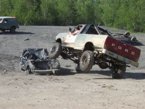 1996 Ford Bronco - A modified Ford truck is shown crushing a car, with another vehicle overturned in the background at an off-road park.