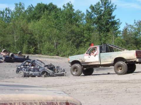 1996 Ford Bronco - Wrecked cars and a lifted pickup truck are visible in a dirt lot with trees in the background.