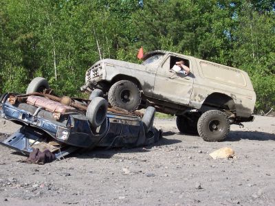 1996 Ford Bronco - A tan SUV is perched atop a severely damaged blue car in a dirt lot, with trees in the background.