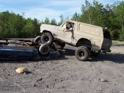 1996 Ford Bronco - A tan Ford Bronco is positioned on top of a flipped blue car in a dirt lot with trees in the background.