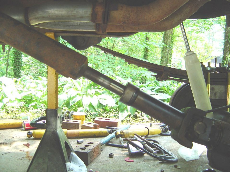 Underside of a raised vehicle showing driveshaft, exhaust, leaf spring and new shock, with tools scattered on ground.
