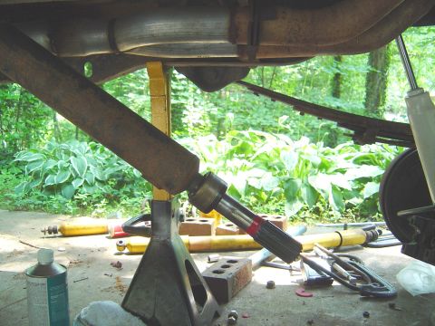 1986 Ford Bronco - Underside view of a vehicle raised on a yellow jack stand, showing the exhaust pipes, driveshaft with slip yoke and splined output shaft, a…