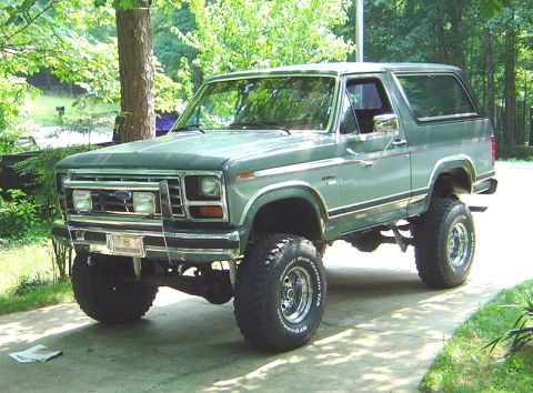 1986 Ford Bronco - A dark green two-door Ford Bronco SUV lifted on large BFGoodrich Mudterrain T/A tires with chrome wheels, parked on a concrete driveway sur…
