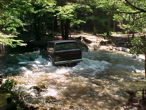 1996 Ford Bronco - A dark-colored Ford Bronco navigates a rocky stream bed surrounded by lush green trees.