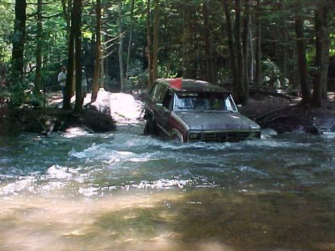 1996 Ford Bronco - A red and silver SUV navigates a shallow stream surrounded by trees.