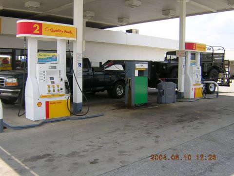 1984 Chevrolet Blazer - A dark pickup truck tows a military-style trailer carrying another vehicle at a gas station.