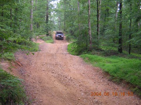 1984 Chevrolet Blazer - A dark colored SUV drives up a gravel road surrounded by dense forest.