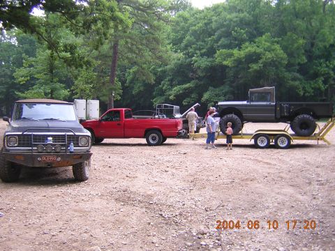 1984 Chevrolet Blazer - A dark off-road vehicle with a prominent brush guard is parked in a gravel lot next to a red pickup truck and a military-style truck on a t…