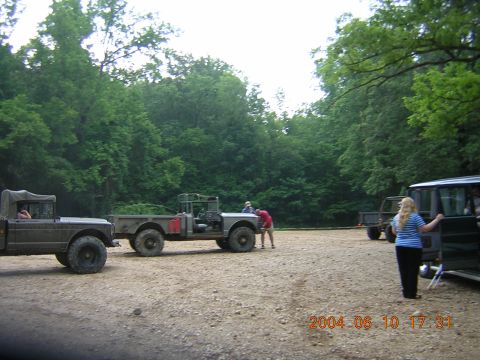 1984 Chevrolet Blazer - Several military-style trucks are parked in a gravel lot surrounded by trees, with people interacting around them.