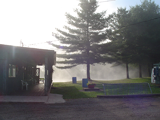 Building with a covered porch and trees in the background with mist. A white vehicle is partially visible on the right